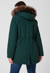 Parka matelassée vert foncé avec capuche doublée de fourrure, cordon de serrage ajustable à la taille et fentes latérales à l'ourlet. Texture de tissu lisse.