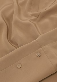 Manche de chemise beige avec deux boutons beiges sur le poignet et des détails de couture visibles sur un tissu lisse.