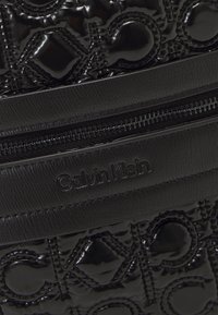 Czarna błyszcząca torba Calvin Klein z wytłoczonym logo, gładką fakturą oraz zamkiem błyskawicznym z poziomym skórzanym paskiem i brandingiem.