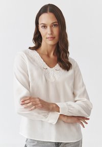 Blusa bianca in tessuto leggero con scollatura a V ricamata, maniche lunghe e polsini abbottonati. Tessuto morbido, vestibilità comoda.