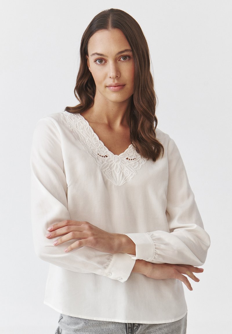 Blusa bianca in tessuto leggero con scollatura a V ricamata, maniche lunghe e polsini abbottonati. Tessuto morbido, vestibilità comoda.