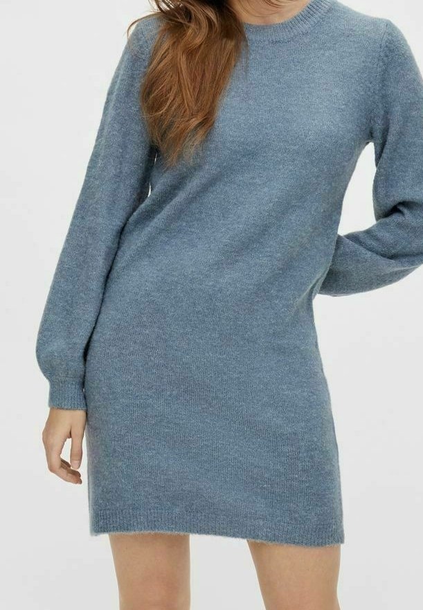 Femme portant une robe-pull en tricot bleu clair à manches longues, debout avec une main sur la hanche devant un fond uni.