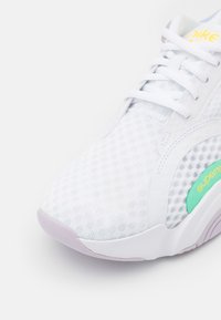 Scarpa sportiva bianca con tomaia in rete, caratterizzata da un accento verde e una suola viola. Design con lacci e imbottitura al tallone.