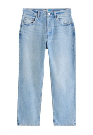 Kvinde står indendørs iført en lyserød polo med korte ærmer, lyseblå jeans og hvide perforerede slip-on sko ved et stort vindue med træer udenfor.
