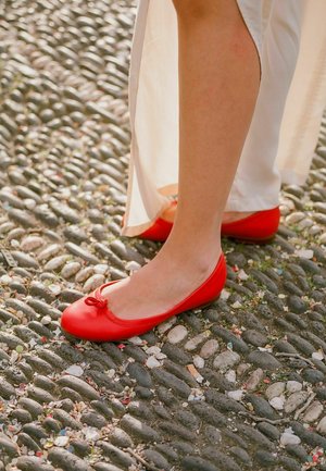 Červené baletní střevíce vyrobené z hladké kůže s kulatou špičkou a malou mašlí vpředu, umístěné na texturovaném žulovém povrchu.