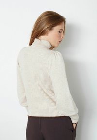 Maglione a collo alto a coste in beige chiaro, con maniche a sbuffo e orlo in contrasto marrone scuro. Tessuto morbido e design aderente.