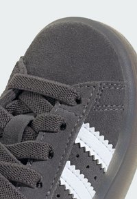 Gray suede sneaker featuring white leather stripes, textured laces, and a rubber sole. Close-up view highlighting stitching and details.