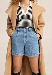 Abrigo color camel, blusa de rayas blanco y negro, y pantalones cortos de denim de tiro alto. El conjunto presenta un corte relajado y costuras en contraste.