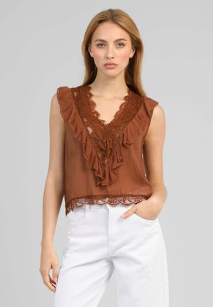 Femme aux longs cheveux blonds portant un chemisier marron sans manches avec dentelle et volants, associé à un pantalon blanc taille haute, debout sur fond gris.