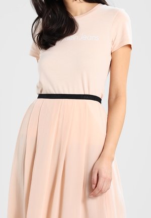 Kvinde iført en lyserød Calvin Klein Jeans t-shirt stoppet ned i et matchende plisseret nederdel med et tyndt sort bælte.