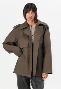 Trench coat supradimensionat de culoare verde măslin, cu un guler structurat, două buzunare pe piept și nasturi aurii, purtat peste un pulover deschis și blugi.