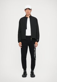 Svart jakke med glidelås foran, hvit T-skjorte under, svarte joggebukser med hvit tekst, og svarte joggesko. Hvit caps fullfører antrekket.