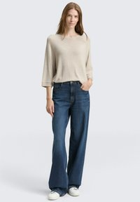 Pullover in maglia beige con maniche ampie abbinato a jeans a gamba larga blu scuro e sneakers bianche; outfit casual dal fit rilassato e dalla texture morbida.