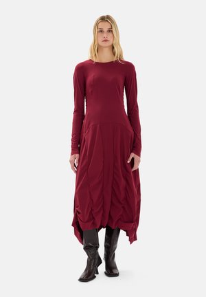 Abito bordeaux a maniche lunghe con texture arricciata, una silhouette svasata e un orlo asimmetrico. Indossato con stivali neri fino al ginocchio.