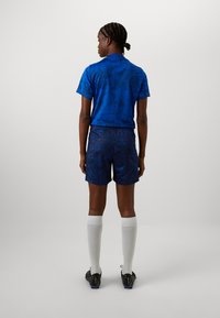 Blue athletic shirt with a textured pattern, paired with navy shorts. Model wears white knee-high socks and black sports shoes.