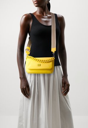 Sac en cuir jaune avec un rabat accentué par une chaîne, une bandoulière ajustable et une texture lisse, porté en bandoulière sur un haut noir et une jupe blanche.