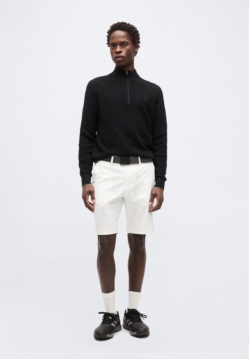 Jeune homme portant un haut noir texturé à manches longues avec fermeture éclair, short blanc, chaussettes blanches, baskets noires, debout contre un fond clair uni.