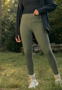 Leggings verde oliva con una cintura alta, textura suave, detalles en la costura lateral, combinados con zapatillas blancas y calcetines crema, en un entorno natural.