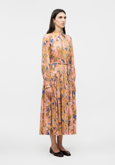 Nő áll hosszú ujjú, gombos virágos ruhában, amely rózsaszín, kék és olívazöld árnyalatú, fekete lapos cipőt visel, egyszerű fehér háttér előtt.