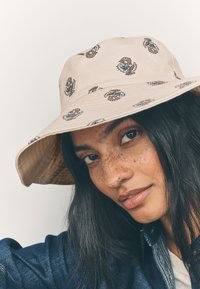 Beiger Fischerhut mit Blumenmustern in Pink, Blau und Schwarz. Hergestellt aus weichem Stoff mit breiter Krempe und strukturierter Innenausstattung.