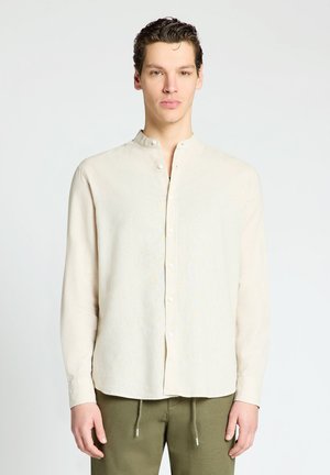 Giovane uomo con capelli corti e scuri che indossa una camicia crema con collo alla mandarino e pantaloni verde oliva, in piedi davanti a uno sfondo bianco semplice.