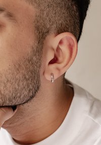 Orecchino a cerchio in argento sul lobo, forma rotonda, texture liscia, design minimale, indossato da una persona con capelli corti e barbuto.