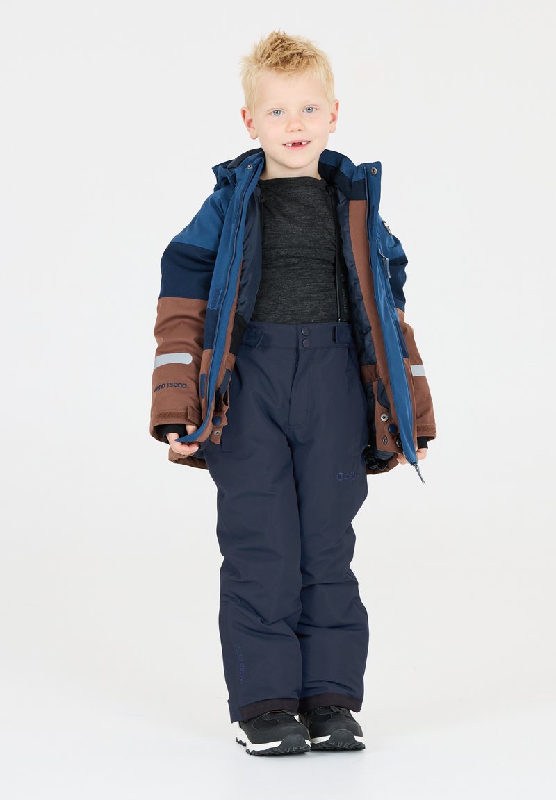 L'ensemble d'hiver pour enfants comprend une veste isolante bleue et marron, un pantalon sombre et des chaussures noires, avec de multiples poches et fermetures éclair.