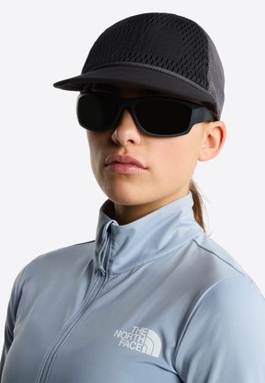 Osoba nosząca czarną siateczkową czapkę, czarne okulary przeciwsłoneczne typu wraparound oraz jasnoniebieską kurtkę na zamek z logo The North Face.