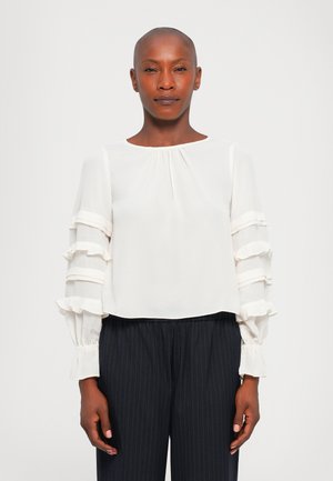 Person with shaved head wearing a white long-sleeve blouse with ruffled sleeves and black pinstripe pants, standing against a plain background.