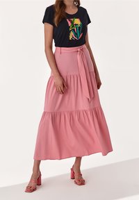 Vrouw in een zwart graphic T-shirt met abstracte vormen, een hooggewaaid roze gelaagd rokje met een gestrikt riem, en roze blokhak sandalen.