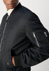 Schwarze Bomberjacke mit glatter Oberfläche, gerippten Bündchen, Reißverschlusstasche am Ärmel und gesticktem Logo auf der Brust.