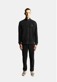 Schwarzer Fleece-Trainingsanzug mit Reißverschlussjacke und passender Hose. Akzentuiert mit einem kleinen gelben Logo. Model steht vor neutralem Hintergrund.