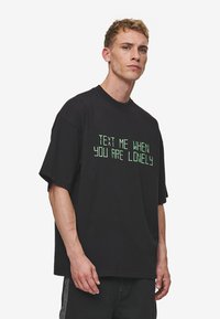 Sort oversized t-shirt lavet af bomuld, med grøn tekst der står "SEND MIG EN BESKED, NÅR DU ER ENSOM" i en blokeret skrifttype.