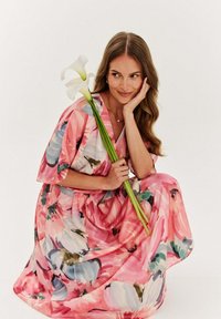 Kvetinové maxi šaty v ružovej farbe s modrými a zelenými akcentmi. Výstrih do V, krátke rukávy a zhromaždený pás. Modelka drží kalu.