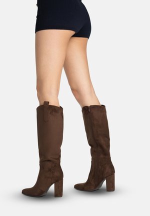 Beine in schwarzen Shorts und hohen braunen Wildlederstiefeln mit Seitenreißverschlüssen, stehend vor einem schlichten hellen Hintergrund.