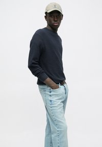 Marineblå crewneck-trøje, lyseblå jeans med lige ben og en beige kasket. Trøjen har ribbede manchetter, og jeansene har en falmet vask.