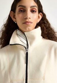 Mladá žena s dlouhými kudrnatými vlasy, která má na sobě náušnice s perlami a krémový fleecový kabátek zapnutý až ke krku přes pruhované tričko.