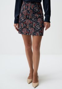 Mini-jupe à motif floral en marine avec des accents rouges et violets, fabriquée en tissu texturé. Associée à des talons aiguilles nude.