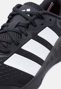 Κοντινό πλάνο μαύρου αθλητικού παπουτσιού με λευκές γεωμετρικές ρίγες, μαύρα κορδόνια και λεπτομέρειες από πλέγμα.