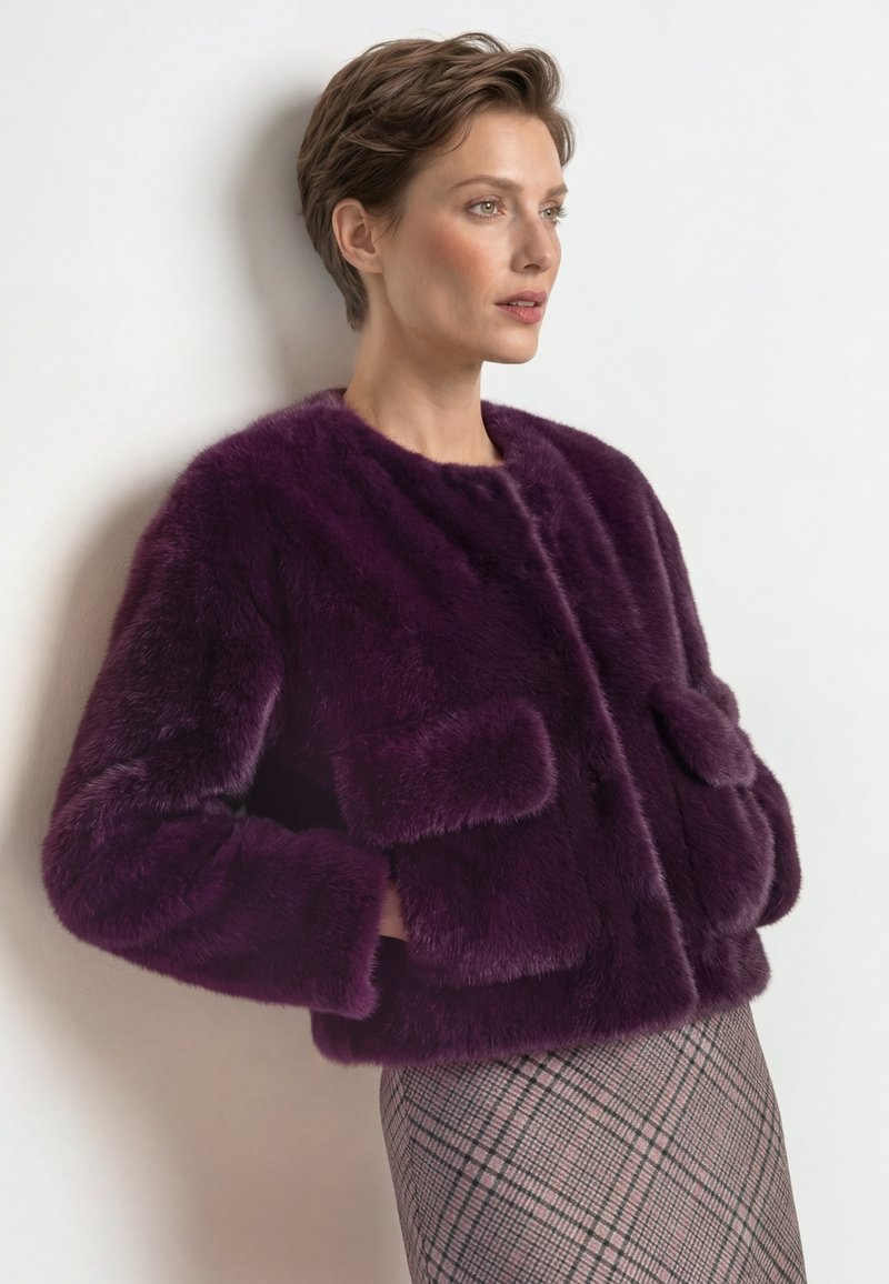 Woman with short brown hair wears a dark purple faux fur jacket and a plaid skirt, standing against a light background.