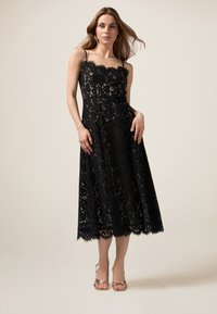 Schwarzes Spitzen-Midi-Kleid mit gewelltem Ausschnitt und Saum. Dünne Träger, halbtransparenter Blumenmuster und fließende Silhouette. Gestylt mit Riemchensandalen.