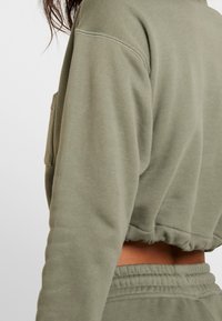 Olivgrön kortad sweatshirt med en mjuk textur och ribbad mudd, med en ficka framtill och elastisk midja, synlig vid fållen.