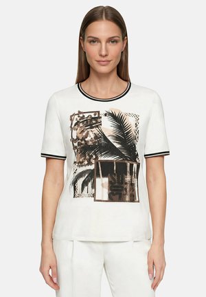 Femme portant un haut blanc à manches courtes avec une bordure noire et un imprimé abstrait de feuilles de palmier et motifs géométriques, associé à un pantalon blanc, debout devant un fond uni.