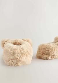 Chaussons en fausse fourrure beige avec un design arrondi, texture douce et petits détails en forme d'oreilles sur les bords. Apparence légère et confortable.