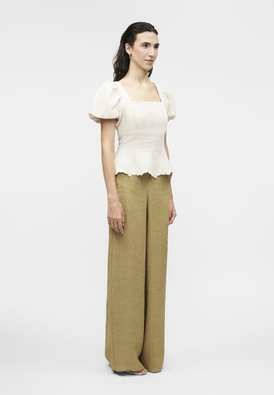 Γυναίκα που στέκεται φορώντας λευκό τοπ με φουσκωτά μανίκια και κυματιστό τελείωμα, μαζί με φαρδιά ελιά πράσινα παντελόνια, σε απλό φόντο.
