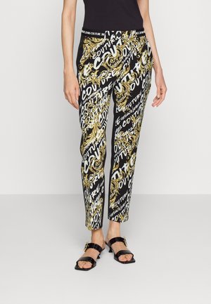 Woman wearing black high-waist pants with gold baroque patterns and white "Couture" text, paired with black heeled sandals.