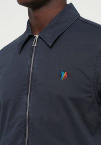 Granatowa kurtka z zamkiem błyskawicznym i spiczastym kołnierzem, z wielokolorowym haftowanym logo konia po lewej stronie klatki piersiowej.