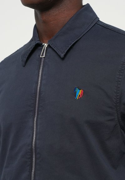 Tamnoplavi sako sa zatvaračem i oštrim ovratnikom, s višebojnim izvezenim logotipom konja na lijevoj strani prsiju.