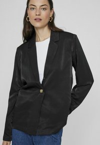 Juodas satino švarkas su vienu auksiniu mygtuku, aštriais atlapais ir ilgomis rankovėmis, derinamas su baltu viršumi ir mėlynomis džinsomis.