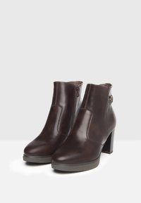 Stivali alla caviglia in pelle marrone con tacco a blocco alto. Presentano una zip laterale e una texture liscia, con punta arrotondata e dettagli minimi.
